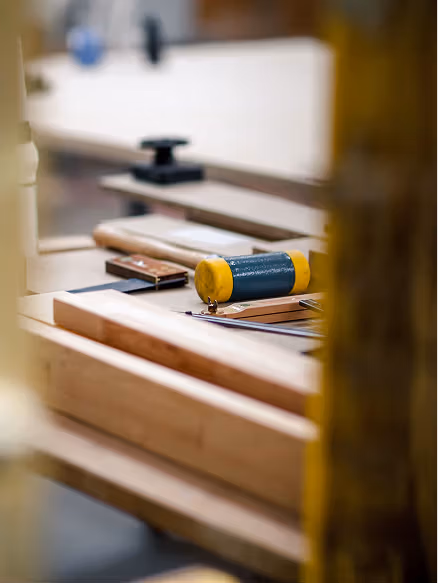 A wooden workbench with various tools and materials, including a yellow pencil, a clamp, and a blurred background.