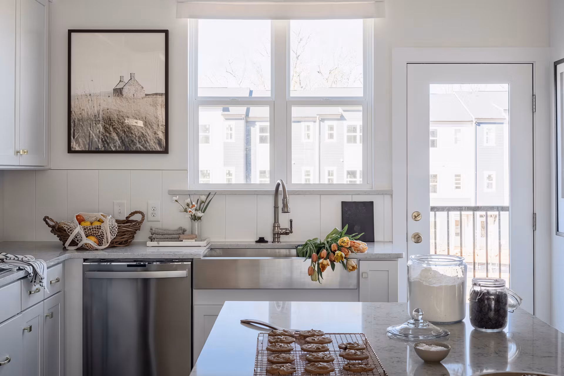 A modern, well-lit kitchen with stainless steel appliances, a farmhouse-style sink, and a framed artwork on the wall, along with various kitchen items and fresh produce on the countertop.